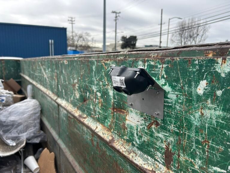 A green dumpster with a sensor mounted on the side used for monitoring or tracking waste levels.