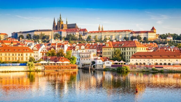 Colorful historic buildings along the Vltava River with Prague Castle rising in the background.