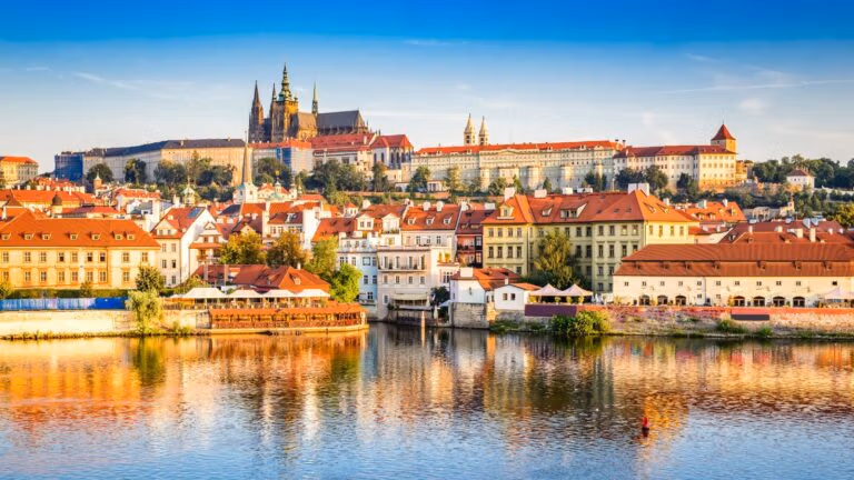 Colorful historic buildings along the Vltava River with Prague Castle rising in the background.