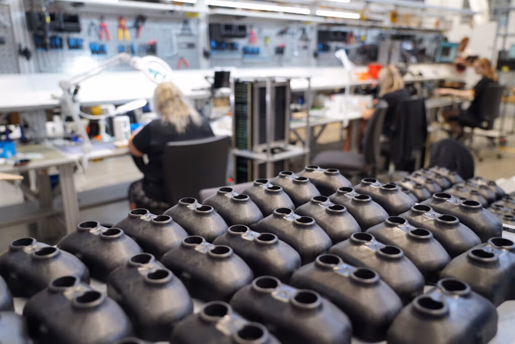 Black plastic components being assembled in a workshop with workers in the background.