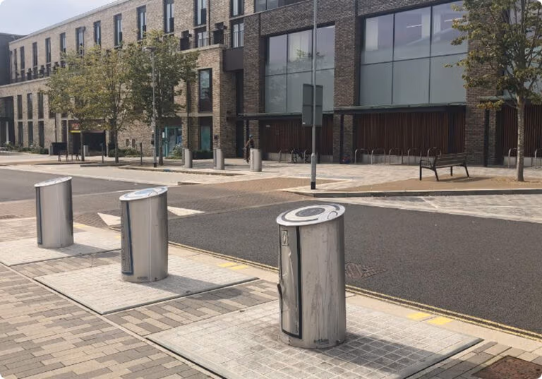 Modern, cylindrical public waste bins installed along a sidewalk in an urban area.