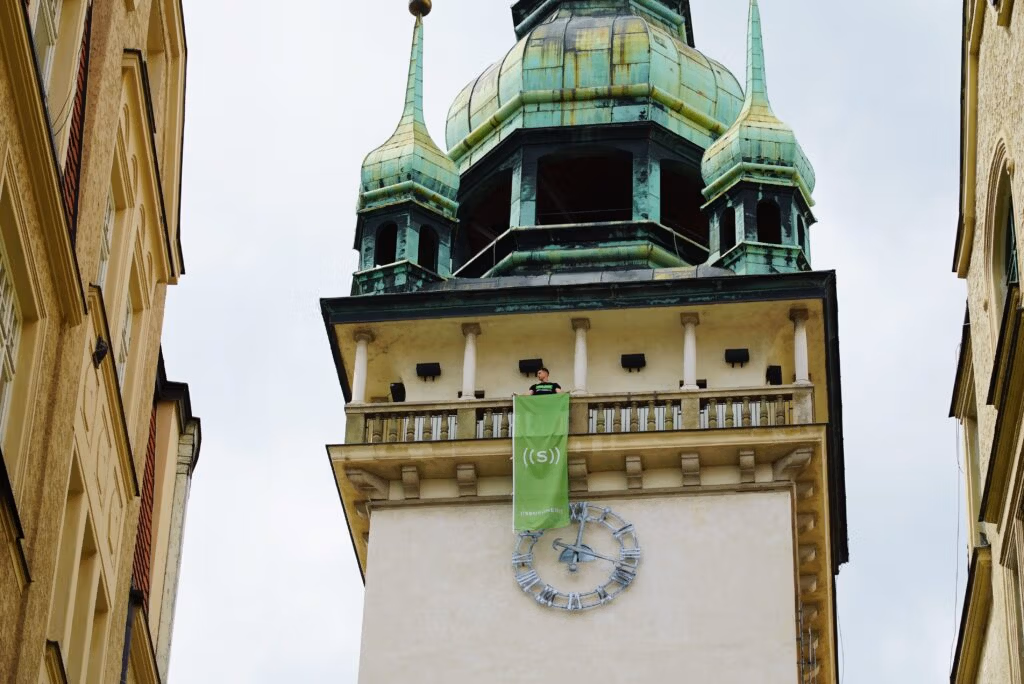 A person hanging a green banner from a tower with a clock in the background.
