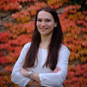 A woman stands confidently with arms crossed, smiling in front of vibrant red foliage.