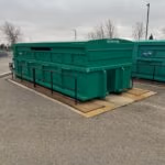 Large green recycling container outdoors, on a concrete pad, enclosed by safety railings.