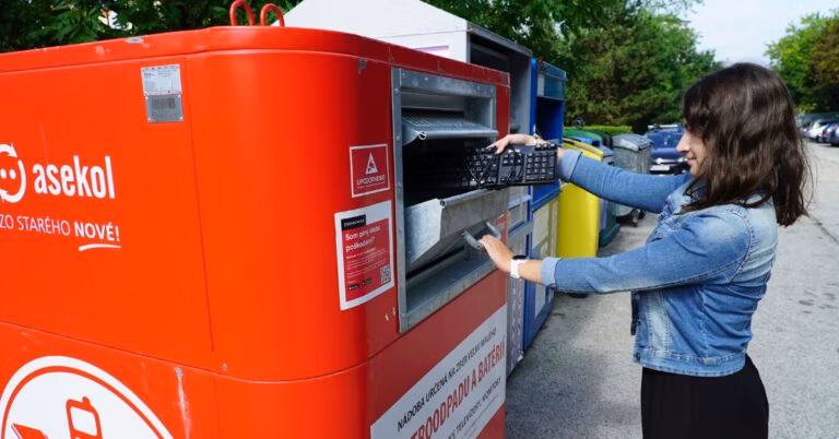 A woman places an old computer keyboard into a large red electronics recycling container outdoors.