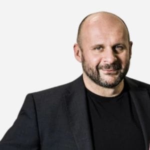 Confident bald man in dark blazer, slight smile, relaxed posture.
