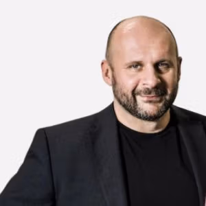 Confident bald man in dark blazer, slight smile, relaxed posture.