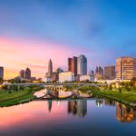 Sunset skyline reflecting in a calm river, with modern buildings and a bridge.