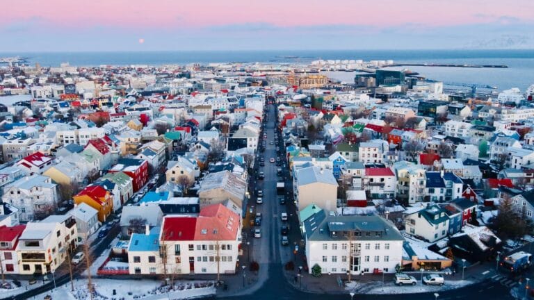 Colorful coastal cityscape with snowy rooftops and mountains, viewed from above at sunset.