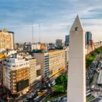 A tall white obelisk rises above a busy city avenue lined with buildings and trees.