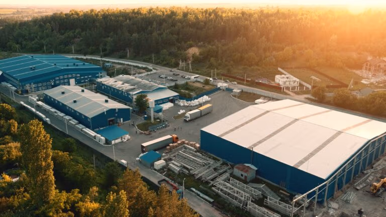 Industrial complex with large blue warehouses surrounded by forest and trucks.
