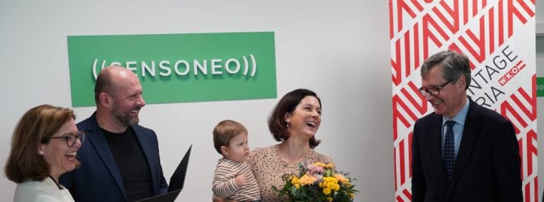 Four adults stand smiling near a “Sensoneo” sign, one woman holds a child and flowers.