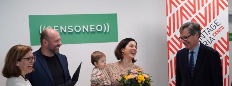 Four adults stand smiling near a “Sensoneo” sign, one woman holds a child and flowers.
