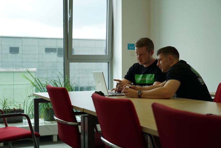Two people at a conference table collaborating on a laptop in a bright office.