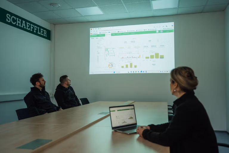 Three colleagues review a projected data dashboard in a meeting room using laptops and charts.