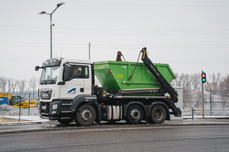 Truck transporting a large green waste container at an industrial site.
