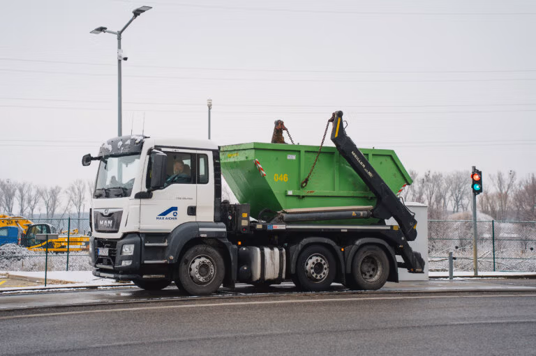 Truck transporting a large green waste container at an industrial site.