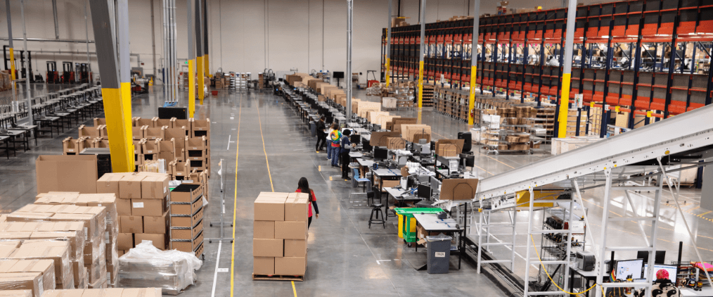 Large warehouse with workers processing packages along assembly lines and stacked shipping boxes.