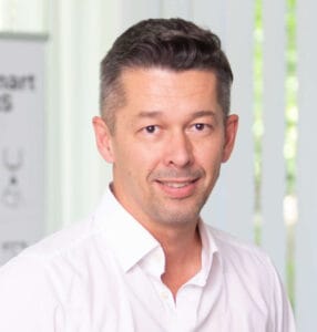 Man in a white shirt smiling in a bright office setting.