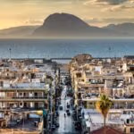 A coastal cityscape at sunset with dense buildings leading toward the sea and mountains.