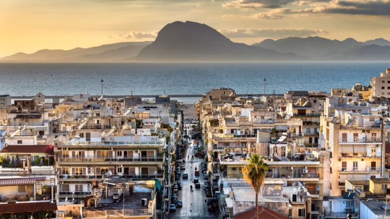 A coastal cityscape at sunset with dense buildings leading toward the sea and mountains.