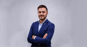 A man in a blue suit stands confidently with arms crossed against a plain background.