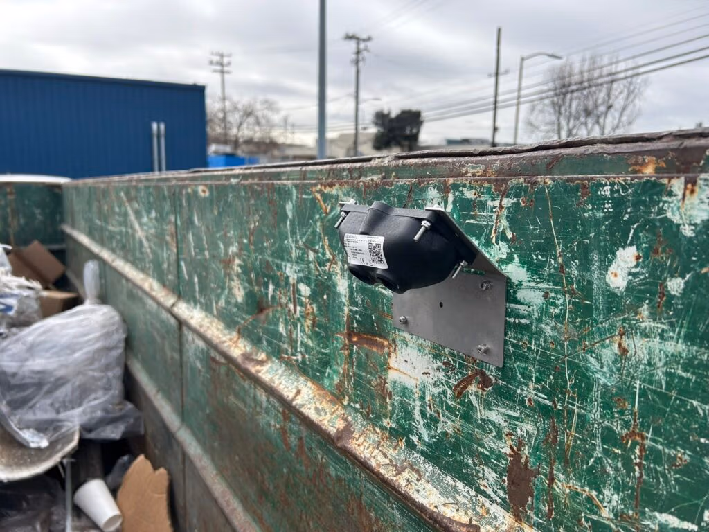 A mounted fill-level sensor attached to a worn, rusty green industrial dumpster wall.