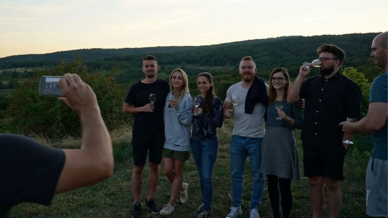 A group of people pose outdoors at sunset, holding wine glasses and smiling together.