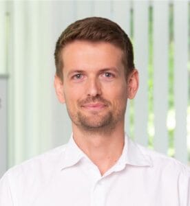 A man in a white shirt looks at the camera, smiling softly with a calm expression.