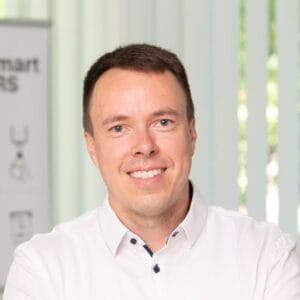 A man in a white shirt smiling, photographed indoors in a bright, professional setting.