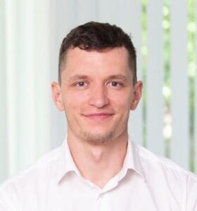 A young man in a white shirt smiles softly while posing indoors near bright vertical blinds.