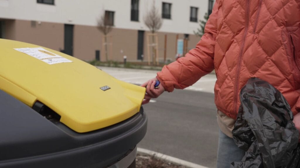 Person opening a recycling bin lid at an outdoor waste collection point.
