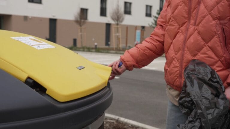 Person opening a recycling bin lid at an outdoor waste collection point.
