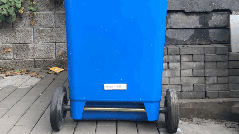 Blue wheeled bin on pavement near a wall and stacked bricks.
