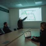 A group of employees in a meeting room reviews a projected facility map on the wall.