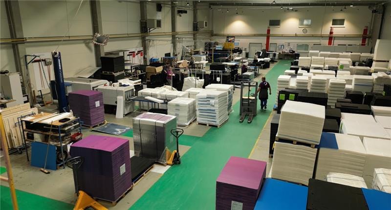 Factory floor with stacked foam materials and workers moving pallets.
