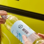 Hands holding a plastic bottle with a deposit barcode before recycling.
