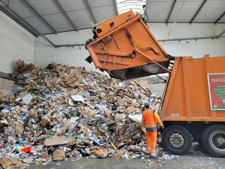 Garbage truck unloading a large pile of cardboard and paper waste indoors.