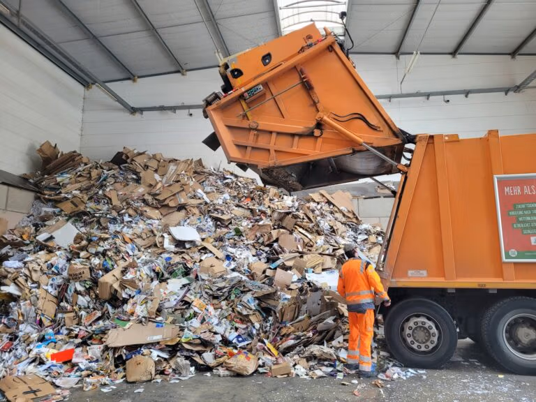 Garbage truck unloading a large pile of cardboard and paper waste indoors.