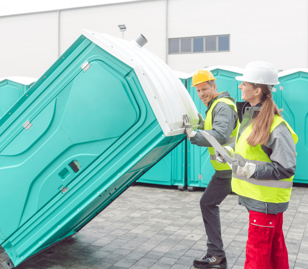 Two workers in safety gear positioning a portable toilet unit.
