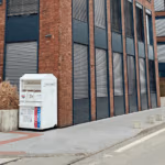 A white clothing-donation bin sits beside a modern brick building on a quiet street.