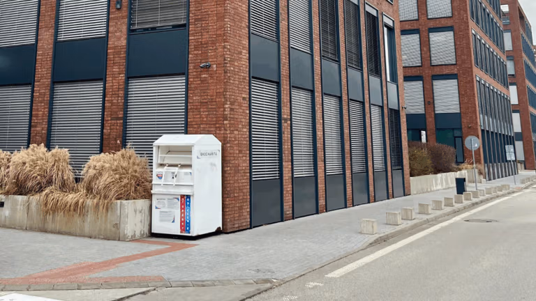 A white clothing-donation bin sits beside a modern brick building on a quiet street.