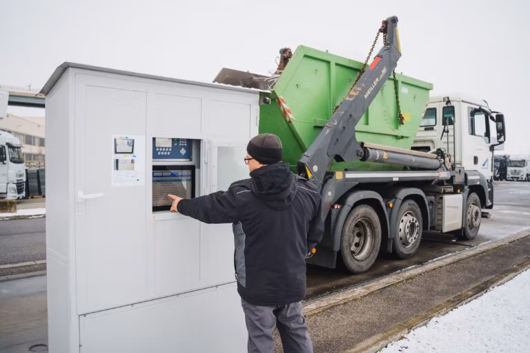 Worker using an outdoor control terminal while a truck lifts a large green container nearby.