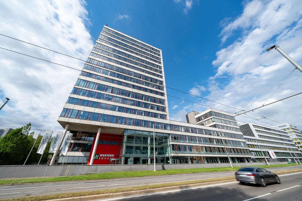 Modern multi-story office building with glass windows, Siemens branding, a main road in front, and a partly cloudy blue sky.